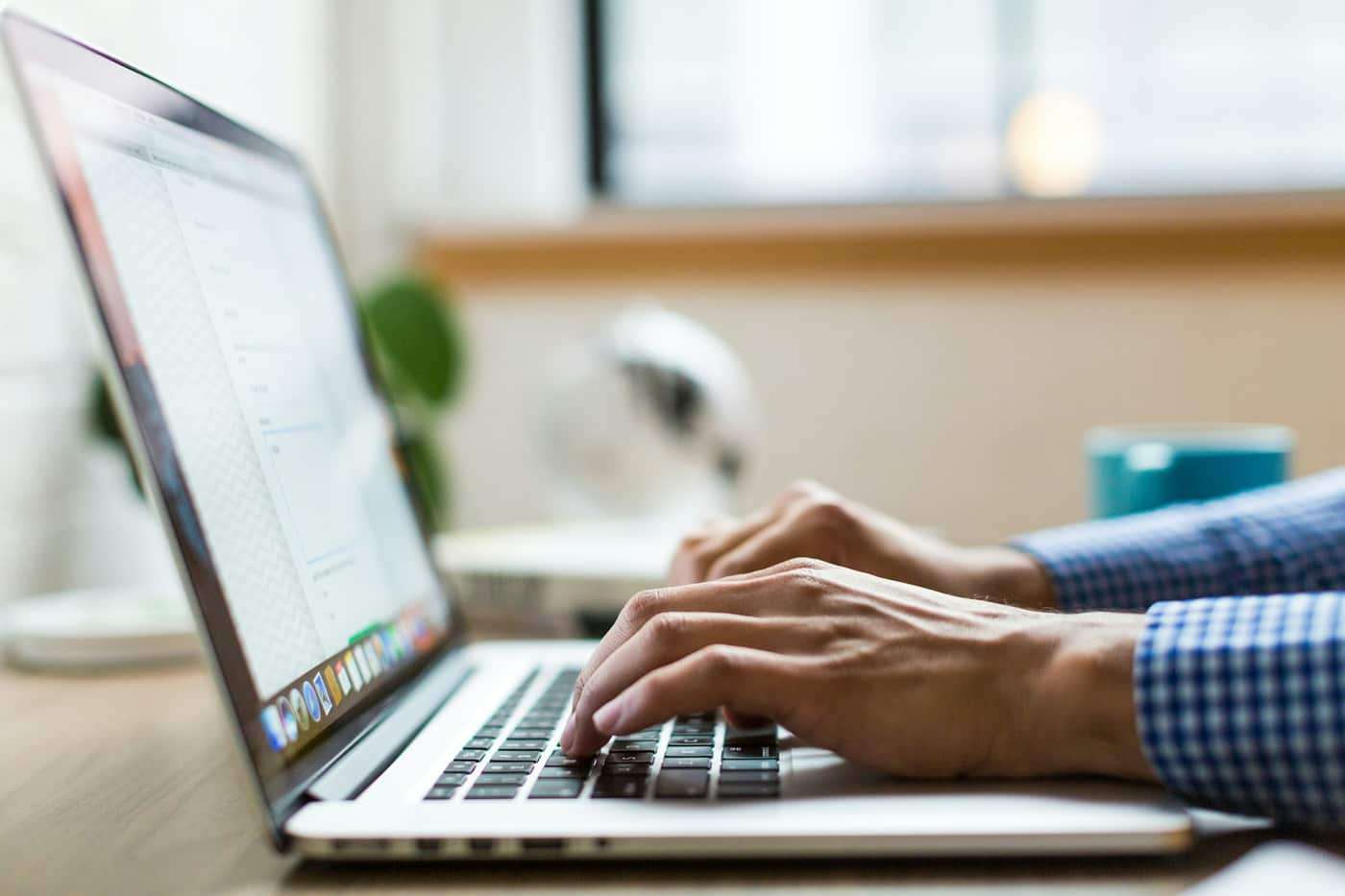 Hands typing on a laptop at a wooden desk by a window.